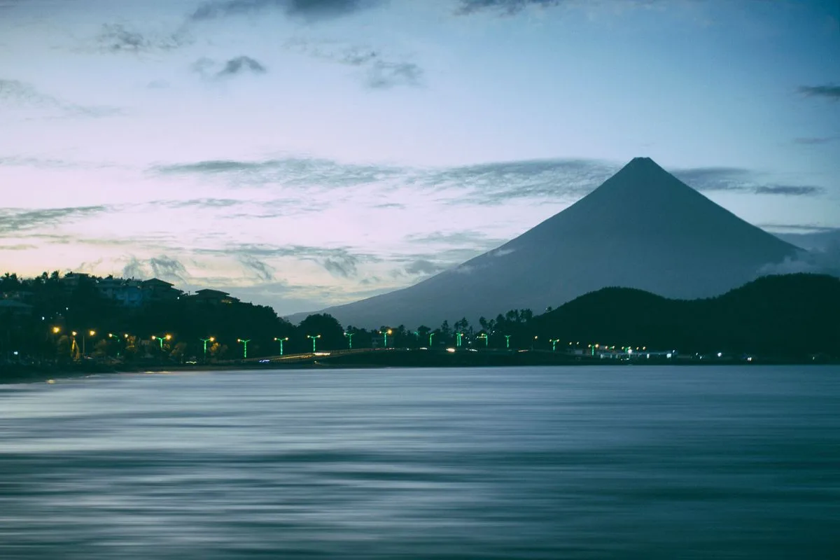 Mayon Volcano at twilight, its silhouette mirrored in calm water under a pale evening sky.